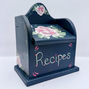 May include: A navy blue wooden recipe box with a floral design. The box features a large pink rose on the lid and the word "Recipes" in white script. The box has a decorative arched top.