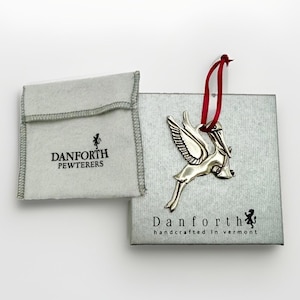 May include: A silver-toned pewter stork ornament with a red ribbon, presented on a square silver box labeled Danforth handcrafted in Vermont, accompanied by a small gray fabric pouch with the text Danforth Pewterers printed in black.