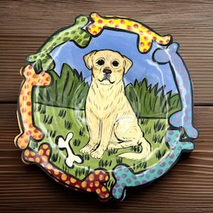 May include: A round, hand-painted ceramic plate featuring a yellow Labrador Retriever sitting in a grassy field. The plate's border is decorated with colorful bone shapes in red, yellow, green, and blue. The background includes a blue sky and green grass.