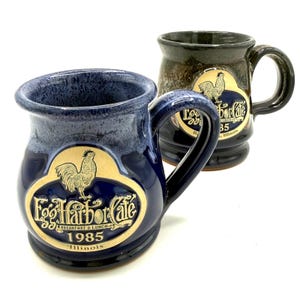 May include: Two ceramic mugs with handles, one in deep blue and one in dark brown, both featuring a speckled glaze finish. Each mug displays a raised emblem with a rooster illustration and the text Egg Harbor Cafe, Breakfast & Lunch, 1985, Illinois.