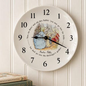 May include: A round white wall clock featuring black numerals and a central illustration of Beatrix Potter's Peter Rabbit characters. The text reads, 'Now, my dears,' said old Mrs. Rabbit, 'you may go into the fields but don't go into Mr. McGregor's garden.'