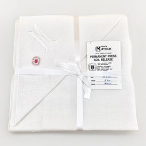 May include: A stack of white linen napkins, tied with a white ribbon. The napkins are embroidered with a letter "M" and a small emblem. A tag reads "MATOUK PERMANENT PRESS SOIL RELEASE" and the size is 17 x 17 inches.