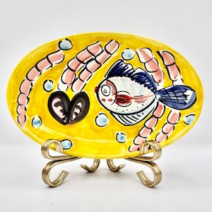 Puede incluir: Un plato ovalado de cerámica pintado a mano con un fondo amarillo brillante. El plato presenta un diseño caprichoso de pez con acentos azules, blancos y rojos, rodeado de formas rosas y negras. Descansa sobre un soporte de metal dorado.