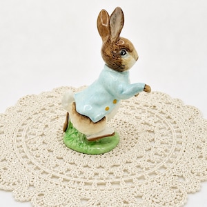 May include: A ceramic figurine of a brown and white rabbit wearing a pale blue jacket with yellow buttons. The rabbit stands on a green base, set against a cream-coloured, crocheted doily.