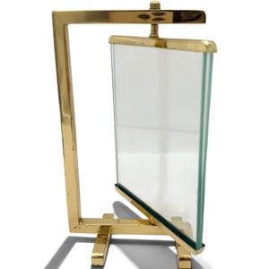May include: A gold-toned metal frame featuring a rectangular glass panel suspended within a rotating structure. The frame has a polished finish and is supported by a base with two horizontal feet, allowing the central glass pane to pivot freely.