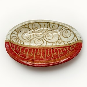 May include: An oval ceramic dish featuring a two-tone design. The upper portion is off-white with a speckled texture and brown circular line patterns. The lower portion is a solid, vibrant red with a repeating pattern of vertical lines and dots.