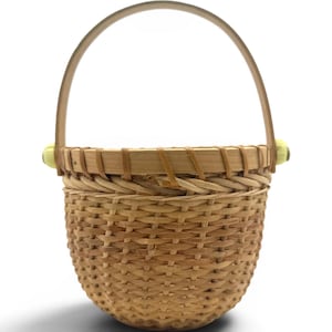 May include: A round woven basket featuring a light brown natural finish with a sturdy arched handle. The basket has a smooth wooden rim and two small, pale yellow decorative knobs where the handle attaches to the main body of the basket.