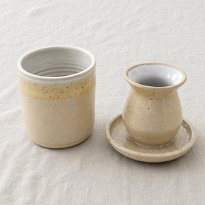 May include: Two ceramic kitchen items. A tall, cylindrical container with a light beige base and a yellow-beige band near the top. Next to it is a butter dish with a lid and a small plate. Both items have a speckled finish.