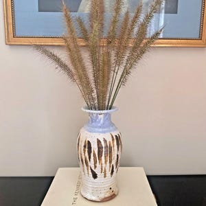 May include: A ceramic vase with a light blue rim and a white body adorned with vertical brown brushstrokes. The vase holds dried ornamental grass. The vase is approximately 25 cm tall.