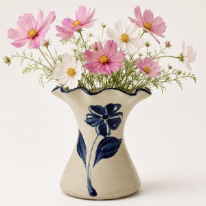 May include: A ceramic vase with a speckled beige finish and a ruffled rim, featuring a hand-painted dark blue flower design on the front. The vase is filled with a bouquet of fresh cosmos flowers in shades of pink and white with yellow centers.