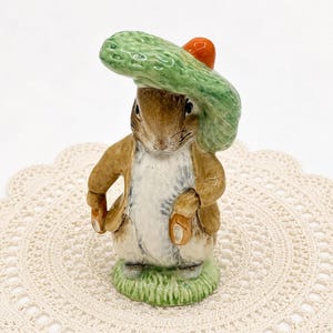 May include: A ceramic rabbit figurine wearing a green hat and a small orange carrot. The rabbit is dressed in a brown jacket and stands on a green grassy base. The figurine has a detailed, hand-painted appearance.