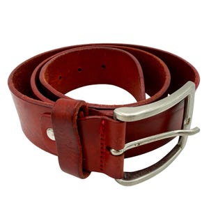 Op de afbeelding: Een opgerolde rode leren riem met een zilverkleurige metalen gesp met één doorn. De riem is voorzien van een bijpassende leren lus en een reeks gestanste gaten langs de riem voor verstelling, met een gladde afwerking.