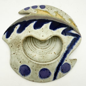 May include: A speckled ceramic dish with an irregular, organic shape. The surface features a cream-colored base with dark blue wave-like patterns, circular spots, and a central recessed area with concentric circular ridges and tan accents.