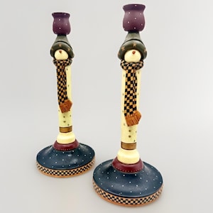 May include: Pair of decorative snowman candle holders. Each holder features a snowman design with a purple top, green hat, checkered scarf, and a navy blue base with white polka dots. The candle holders are approximately 10 inches tall.