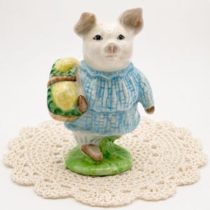May include: A ceramic pig figurine wearing a blue sweater and carrying a basket of yellow fruit. The pig stands on a green base, and the figurine is placed on a cream crocheted doily. The figurine is a charming decorative item.