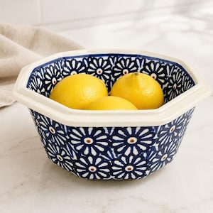 May include: An octagonal ceramic bowl featuring a dark blue and white floral pattern with three bright yellow lemons inside. The bowl has a wide, cream-colored rim and a repeating daisy-like motif covering both the interior and exterior surfaces.