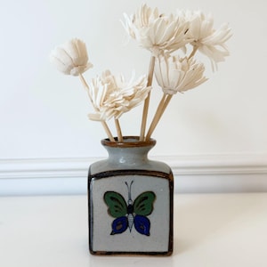 May include: A small, square ceramic vase with a butterfly design in green and blue. The vase is filled with several white, dried flower stems. The vase has a grey background with brown trim.