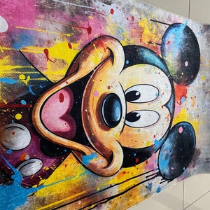 May include: A rectangular area rug featuring a large, centered illustration of Mickey Mouse. The design incorporates a vibrant, multi-colored paint splatter effect with splashes of yellow, pink, blue, and black across a textured, neutral-toned background.