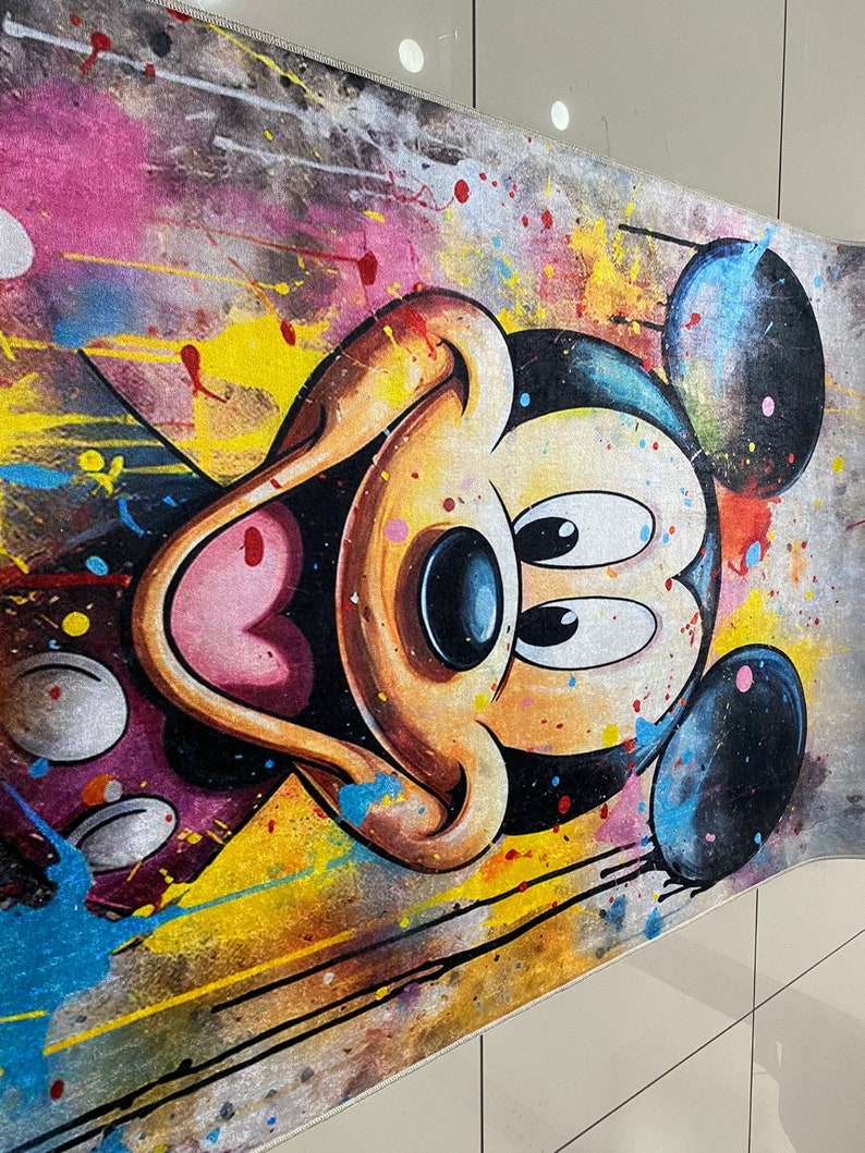 May include: A rectangular area rug featuring a large, centered illustration of Mickey Mouse. The design incorporates a vibrant, multi-colored paint splatter effect with splashes of yellow, pink, blue, and black across a textured, neutral-toned background.