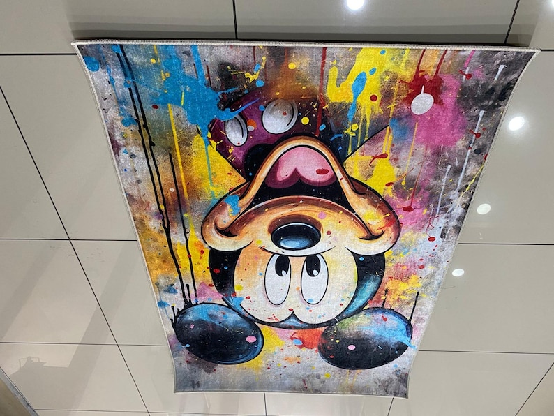 K&ouml;nnte beinhalten: Ein rechteckiger Teppich mit einem auf dem Kopf stehenden Cartoon-Mausgesicht mit gro&szlig;en runden Ohren. Das Design zeigt einen lebendigen Farbspritzer-Effekt in Gelb, Blau, Pink und Schwarz auf einem strukturierten, abgenutzten Hintergrund.