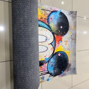 May include: A rectangular area rug featuring a stylized graphic of a mouse head with large black ears. The design incorporates vibrant paint splatters in yellow, blue, and red against a neutral background. The underside has a dark gray textured backing.