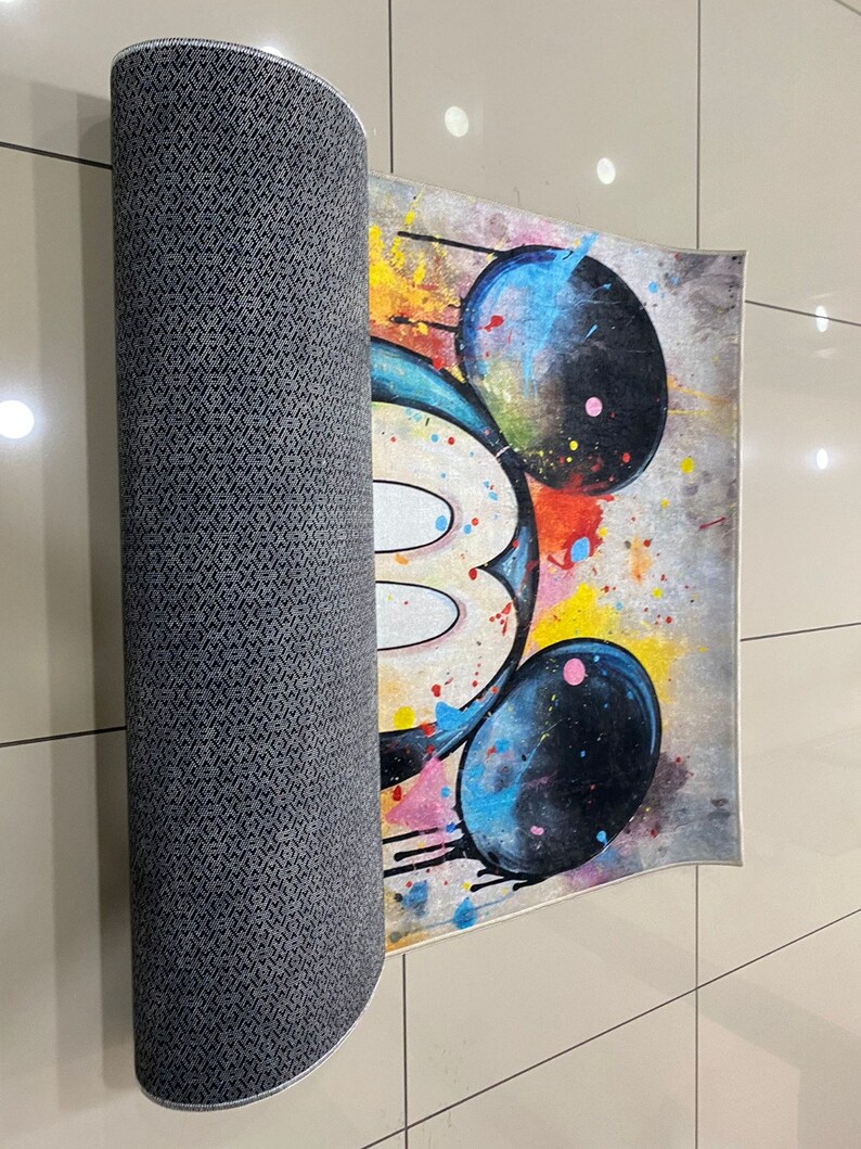 May include: A rectangular area rug featuring a stylized graphic of a mouse head with large black ears. The design incorporates vibrant paint splatters in yellow, blue, and red against a neutral background. The underside has a dark gray textured backing.