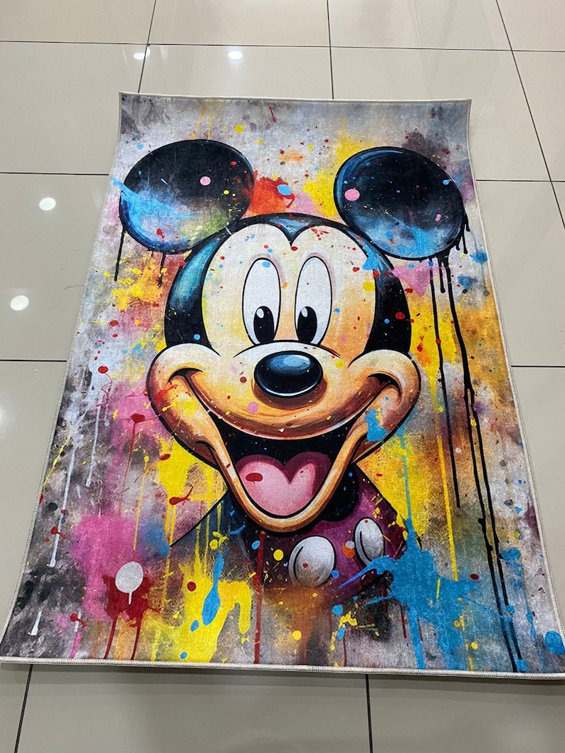 May include: A rectangular area rug featuring a large, centered illustration of Mickey Mouse. The design incorporates a vibrant, multi-colored paint splatter effect with drips of black, yellow, blue, and pink against a textured gray background.