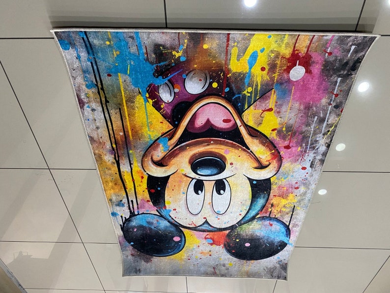 May include: A rectangular area rug featuring an upside-down Mickey Mouse character graphic. The design incorporates a vibrant paint splatter effect with prominent splashes of yellow, blue, pink, and black against a textured, distressed background.