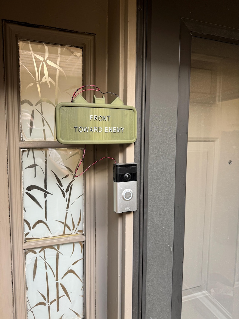 Claymore-style novelty doorbell prank sign installed beside a video doorbell
