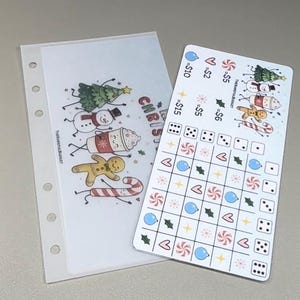 May include: A translucent A6 binder insert with six holes and a Christmas-themed graphic featuring a tree, snowman, cocoa, gingerbread man, and candy cane. Beside it is a white savings tracker card with a grid, dice icons, and holiday symbols.