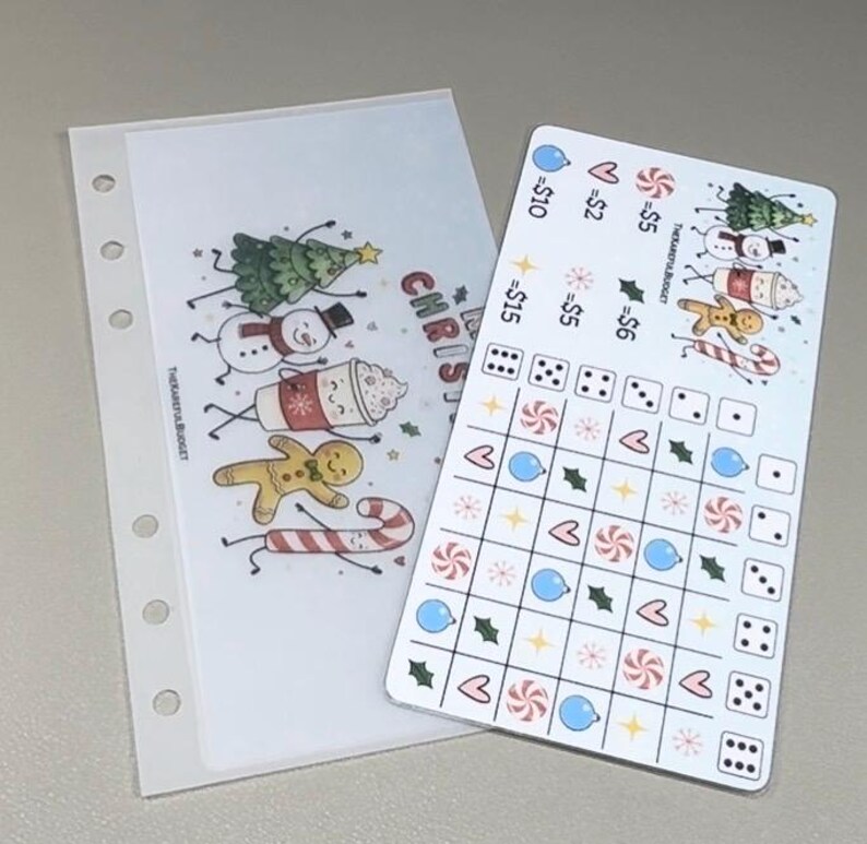 May include: A translucent A6 binder insert with six holes and a Christmas-themed graphic featuring a tree, snowman, cocoa, gingerbread man, and candy cane. Beside it is a white savings tracker card with a grid, dice icons, and holiday symbols.