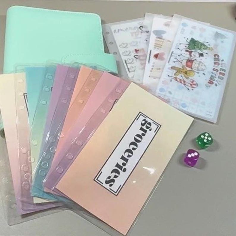 Puede incluir: Una carpeta verde menta con un juego de sobres de pl&aacute;stico transl&uacute;cido multicolor, incluyendo uno etiquetado como Groceries y otro con el texto Merry Christmas y gr&aacute;ficos navide&ntilde;os, junto a un dado verde y uno morado de seis caras.