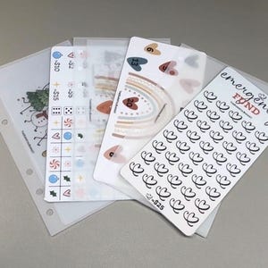 May include: A collection of translucent planner inserts and sticker sheets featuring various designs including a rainbow, hearts, and a tracker labeled emergency fund with a heart icon and the text = $25, alongside icons for $2, $5, $10, and dice.