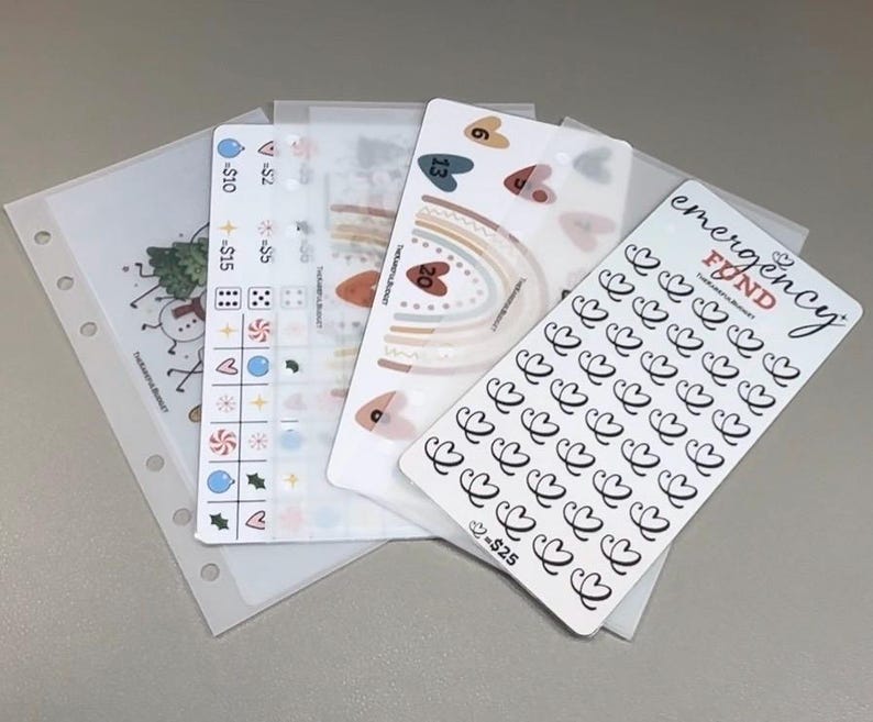 May include: A collection of translucent planner inserts and sticker sheets featuring various designs including a rainbow, hearts, and a tracker labeled emergency fund with a heart icon and the text = $25, alongside icons for $2, $5, $10, and dice.