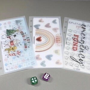 May include: Three rectangular, translucent plastic cash envelopes with punched holes for binders. One features Christmas characters and text, one a rainbow with hearts and the text DEBT FREE, and one the text EMERGENCY FUND with heart icons. Two dice are included.
