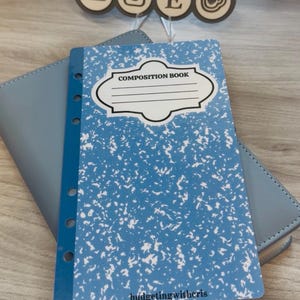 May include: A blue composition book with a white speckled design and the words "COMPOSITION BOOK" in a white label. The book is on top of a blue wallet. The text "budgetingwithcris" is at the bottom.