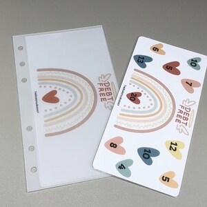 May include: A clear plastic binder pocket with a rainbow and the text DEBT FREE next to a white card featuring a rainbow, the text DEBT FREE, and several colored hearts labeled with numbers including 4, 5, 6, 7, 8, 10, 12, 13, and 20.