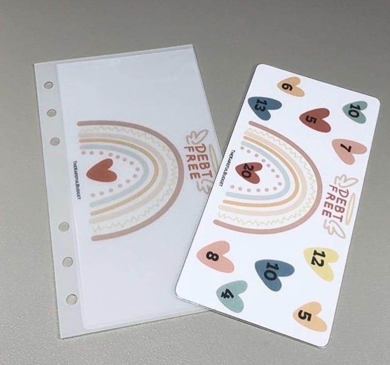 May include: A clear plastic binder pocket with a rainbow and the text DEBT FREE next to a white card featuring a rainbow, the text DEBT FREE, and several colored hearts labeled with numbers including 4, 5, 6, 7, 8, 10, 12, 13, and 20.