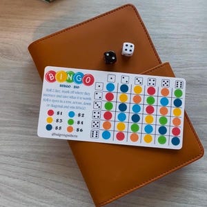Könnte beinhalten: Ein braunes Leder-Notizbuch mit einer Bingo-Karte und zwei Würfeln. Die Bingo-Karte hat bunte Kreise und den Text "BINGO". Die Würfel sind schwarz und weiß.