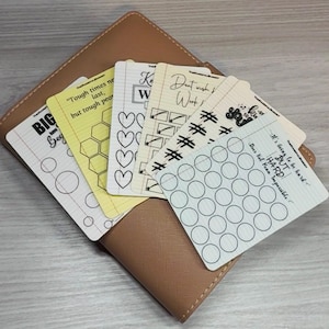 Könnte beinhalten: Ein Set aus fünf rechteckigen Gewohnheits-Tracker-Karten mit liniertem Papierhintergrund, die verschiedene schwarze Tintenformen wie Kreise, Herzen und Sechsecke zur Fortschrittsverfolgung zeigen, platziert auf einem beigen Notizbuchumschlag.