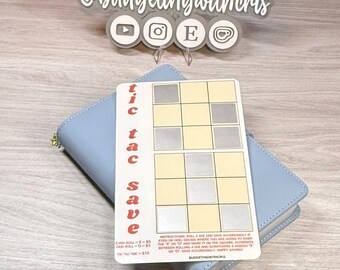 May include: A light blue wallet with a gold zipper holds a "Tic Tac Save" card. The card has a tic-tac-toe grid with instructions and the text "@budgetingwithcris". The card is for saving money.