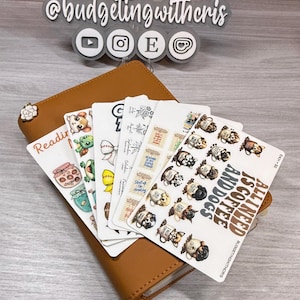 Può includere: Un portafoglio in pelle marrone con un accento floreale contiene diversi fogli di adesivi. Gli adesivi presentano vari disegni, tra cui animali dei cartoni animati, tazze di caffè e il testo "ALL I NEED IS COFFEE AND DOGS."