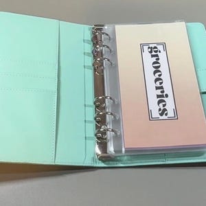 Puede incluir: Una carpeta de seis anillas en color verde menta con cierre magn&eacute;tico. El interior incluye varias ranuras para tarjetas en el lado izquierdo y una funda de pl&aacute;stico transparente a la derecha con una tarjeta que tiene el texto groceries.