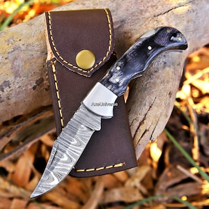 May include: A folding pocket knife with a Damascus steel blade featuring a wavy pattern and a dark, marbled handle. The knife includes a silver bolster and is positioned on a brown leather sheath with yellow stitching and a gold-coloured snap button.