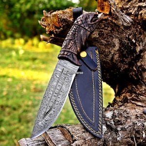 May include: A fixed-blade knife with a patterned Damascus steel blade and a dark brown carved wooden handle featuring an eagle head design. The knife is accompanied by a black leather sheath with yellow stitching and a gold-coloured snap button.