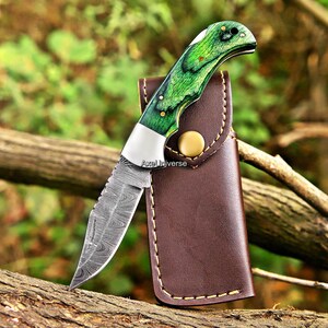 May include: A folding pocket knife with a patterned Damascus steel blade and a bright green wood handle with metal rivets. The knife is positioned in front of a brown leather sheath featuring a gold-coloured snap button and visible stitching.