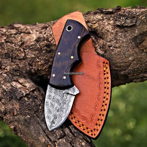 May include: A fixed-blade knife with a dark wood handle featuring brass pins and a patterned Damascus steel blade. The knife is accompanied by a tan leather sheath with decorative stitching and the text AxeUniverse printed on the blade.