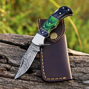 May include: A folding pocket knife with a Damascus steel blade featuring a wavy pattern, a handle made of black and green wood with silver pins, and a silver bolster. The knife is accompanied by a brown leather sheath with yellow stitching.