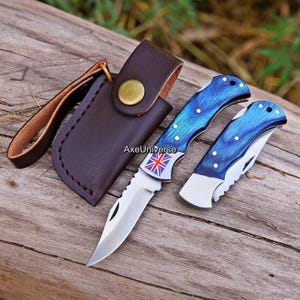 May include: Two folding pocket knives with blue wood-grain handles and silver metal blades. One knife is open, displaying a Union Jack flag graphic near the bolster. A dark brown leather sheath with a brass snap closure is included in the set.