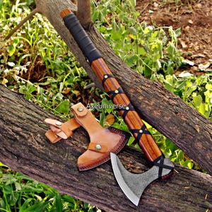 May include: Axe with a wooden handle and a metal blade, resting on a log. The handle is wrapped in black leather and secured with black straps and brass rivets. A leather sheath protects the blade. The text "AxeUniverse" is visible.